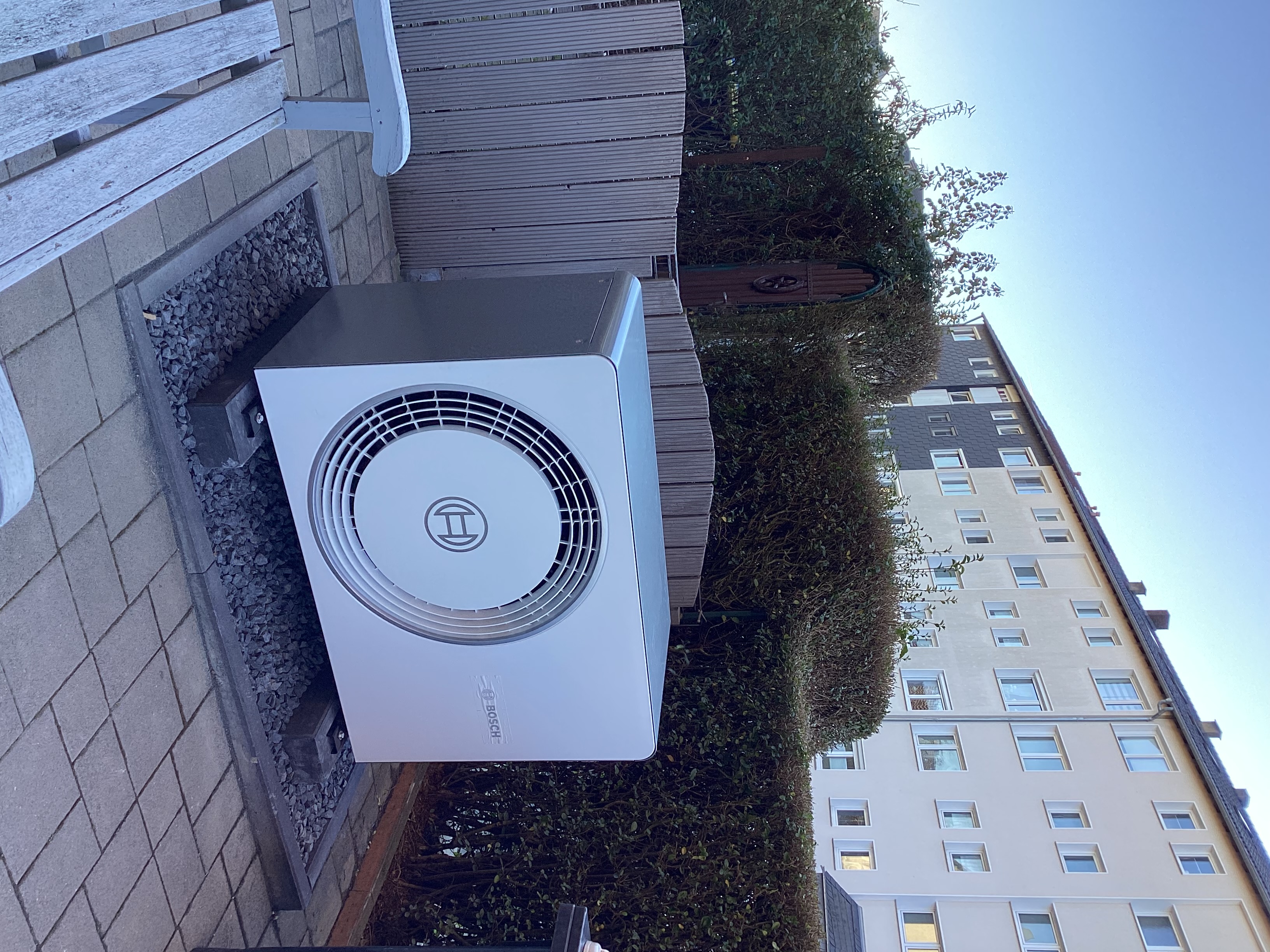 Eine weiße Bosch-Wärmepumpe steht auf einem Kiesbett neben einer Holzbank und ist von Hecken und Sichtschutz umgeben.